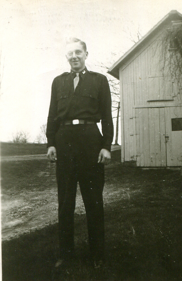 man in dark clothing standing in front of a barn with sun shining behind him