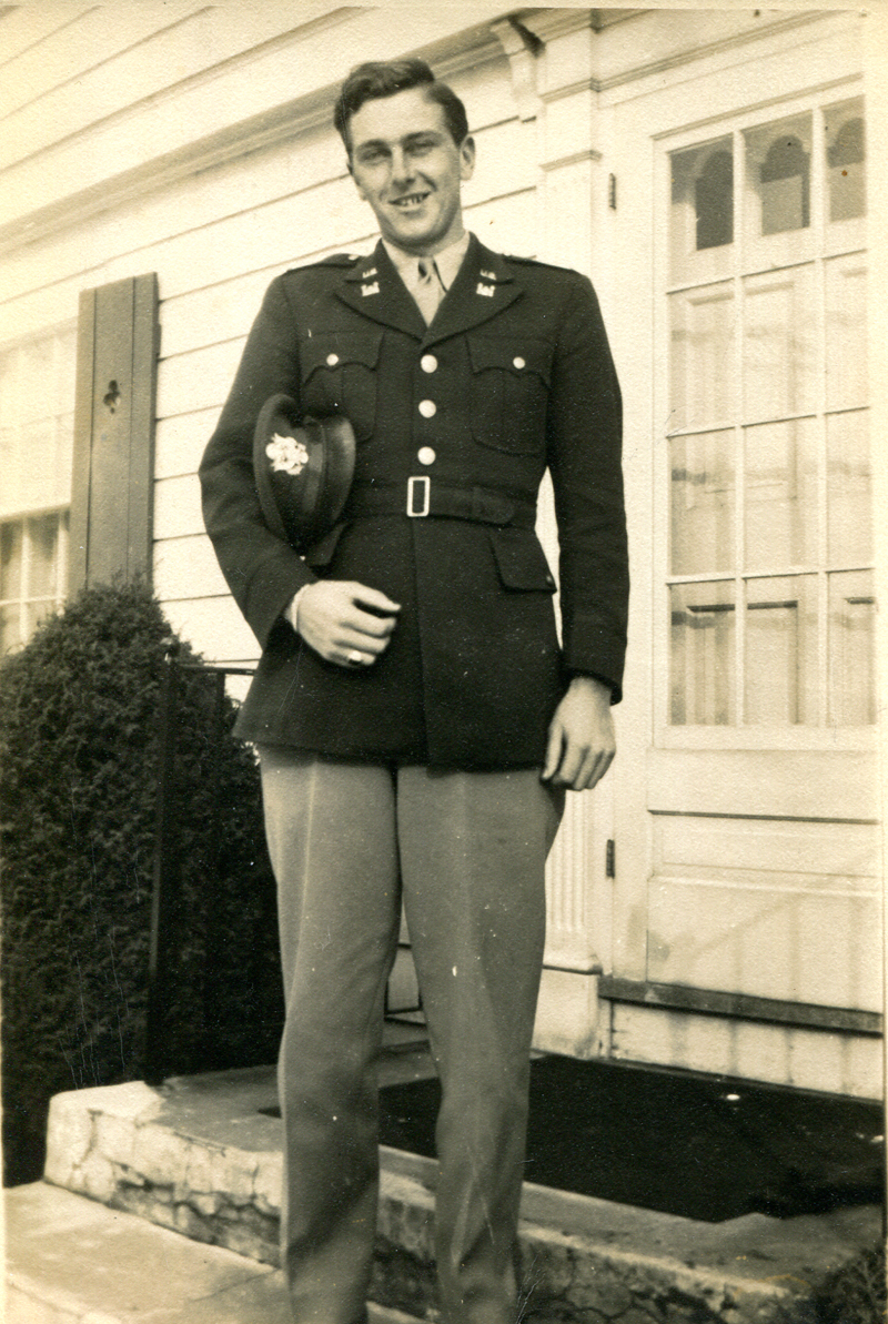 man in uniform with a helmet under his right arm in front of a house