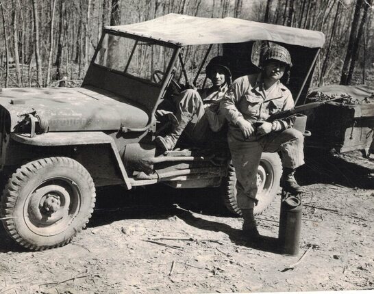 two uniformed men with jeep, one sitting inside it and one standing near it with a rifle
