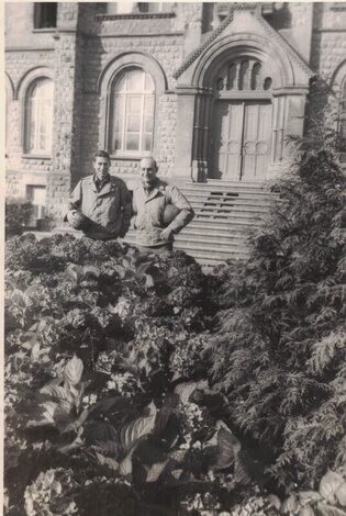 two men standing behind some bushes and in front of a stone building