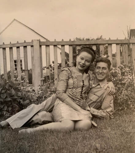 man in uniform and woman sitting on the grass in front of a fence and flowers